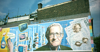 A photograph shows a mural on the side of a building. The mural includes Chomsky's face, along with some newspapers, televisi