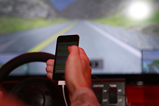 A person's right hand is holding a cellular phone. The person is in the driver's seat of an automobile while on the road.