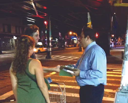 An image of one person holding a clipboard, shaking hands with another person. A third person stands nearby.