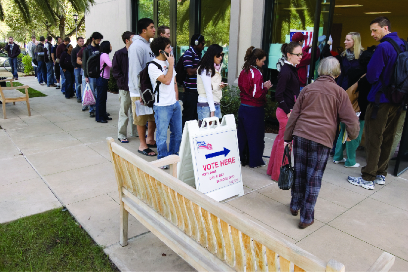 An image of several people standing in line outside of a building. A sign near the front of the line and the building entranc