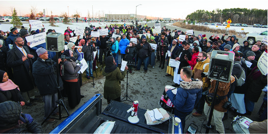 A group of people gathered outside, many with signs, listening to a person speaking.