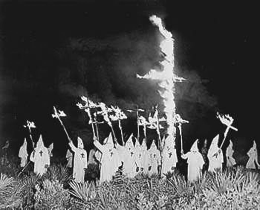 A photo of a group of people wearing robes and pointed hats, surrounding a large cross in the ground that is on fire. Several