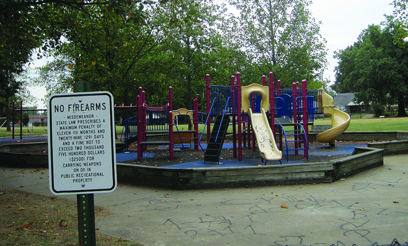 A photo of a sign that reads "No Firearms". A playground can be seen in the background.