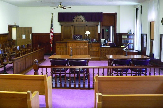 A photo of a typical courtroom, empty of people. In the foreground are benches for attendees, then two tables in the center f