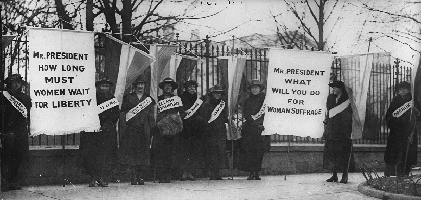 An image of several people standing in front of a fence. Some people are holding banners. The banners read "Mr. President how