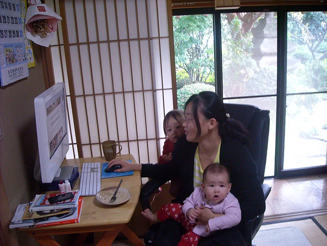 A person sits at a desk while working at a computer, while holding a baby of about six months old. A second child leans again