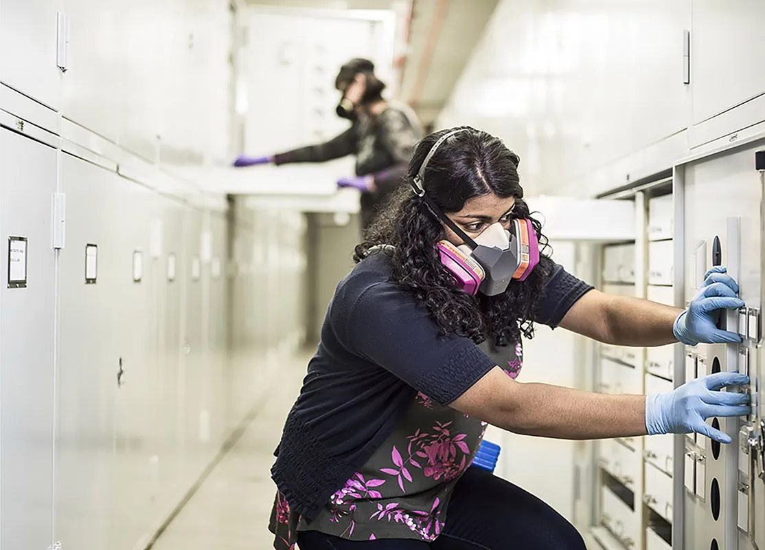 An engineer wearing a large filtration mask and rubber gloves closes a door to a storage unit.