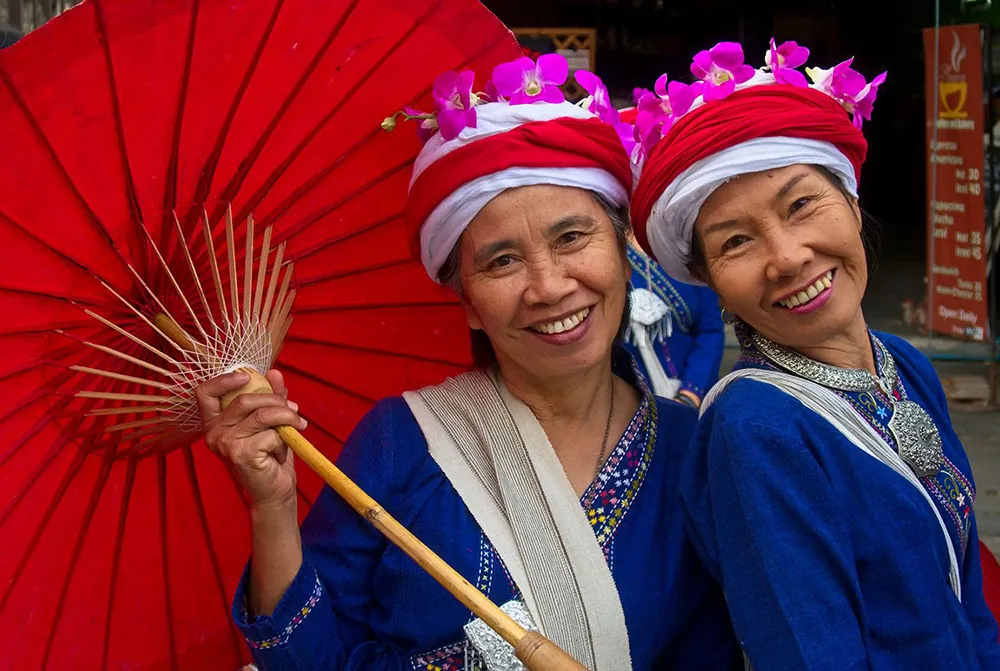 Two people wearing traditional dresses or robes and flowered head coverings stand together. One holds a traditional umbrella.