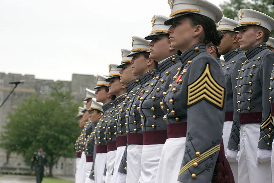 A group of West Point cadets in full dress gray uniforms with white peaked caps. Each wears a red sash around their waist, in