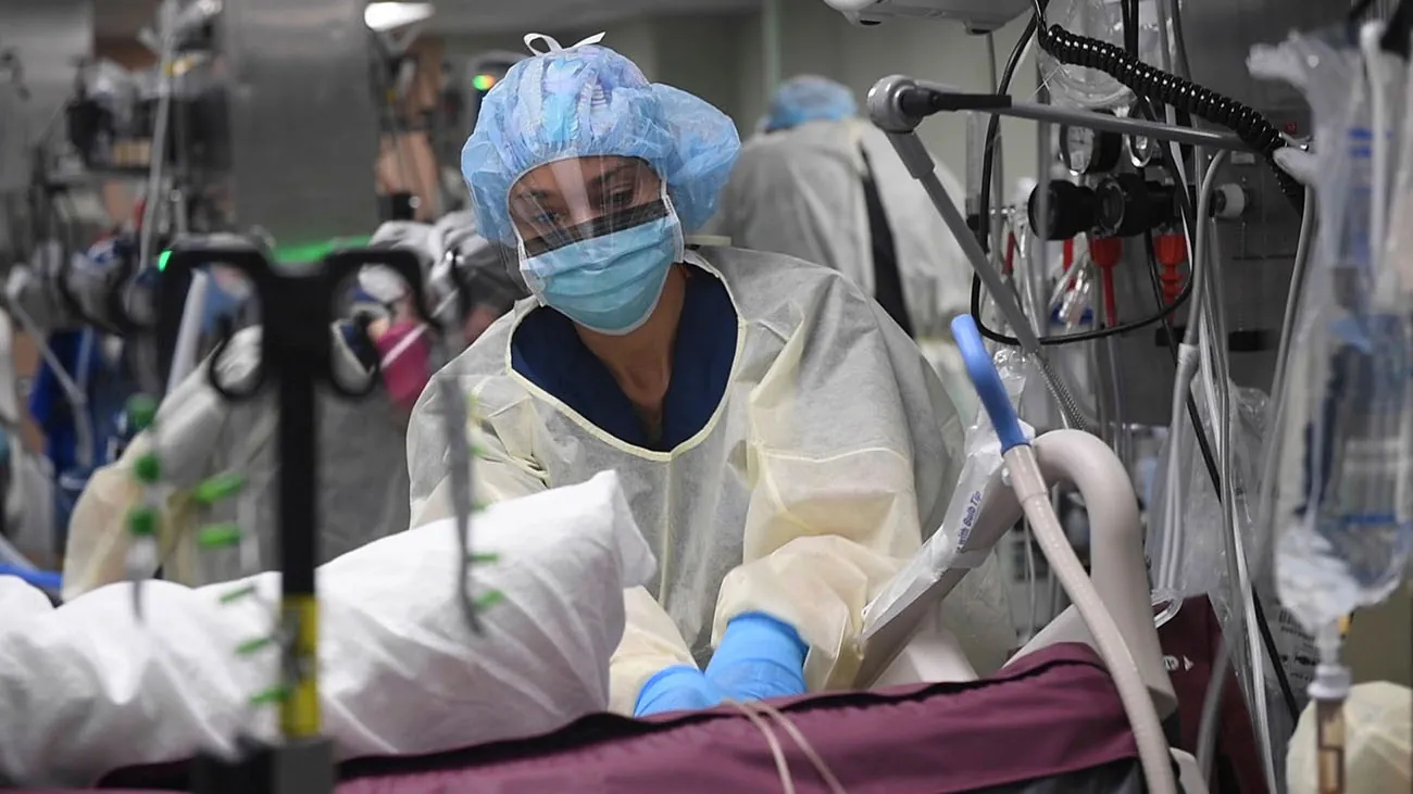 A person in personal protective gear including a mask and face shield works on a hospital bed with extensive equipment.