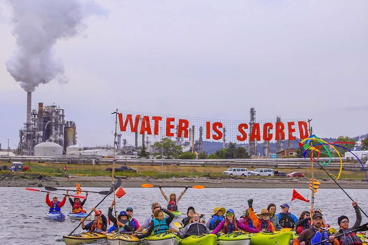 About 20 people in kayaks float in front of an oil refinery. They hold a sign that reads "Water Is Sacred".