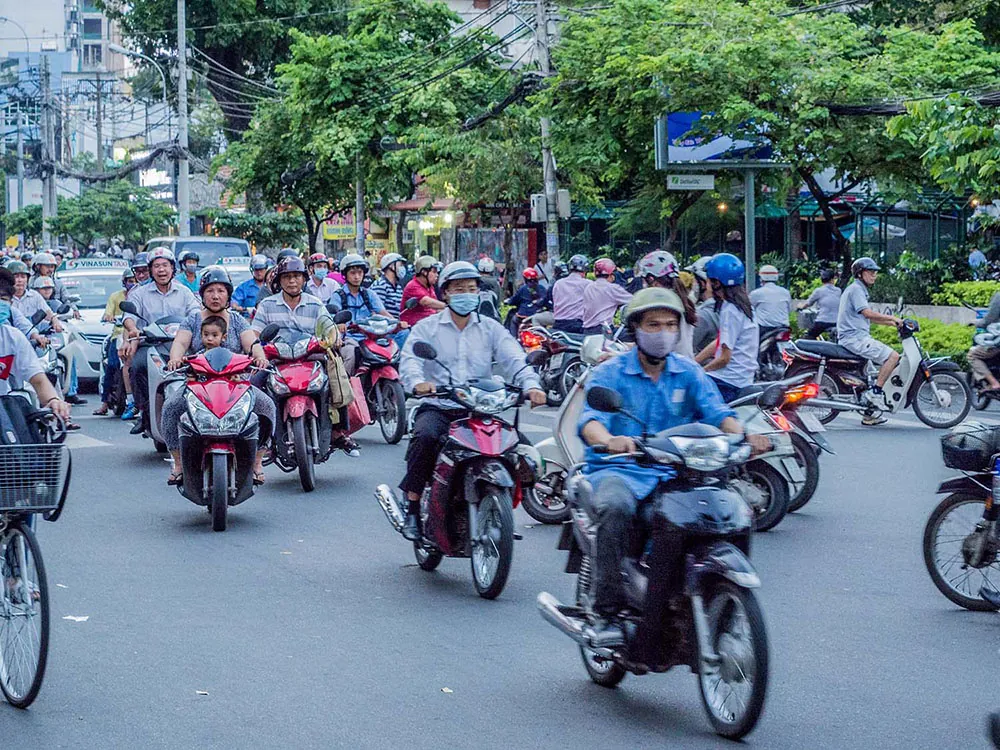 About twenty people on motorcycles and scooters drive along a crowded road with cars and bicycles. Some wear masks and most w