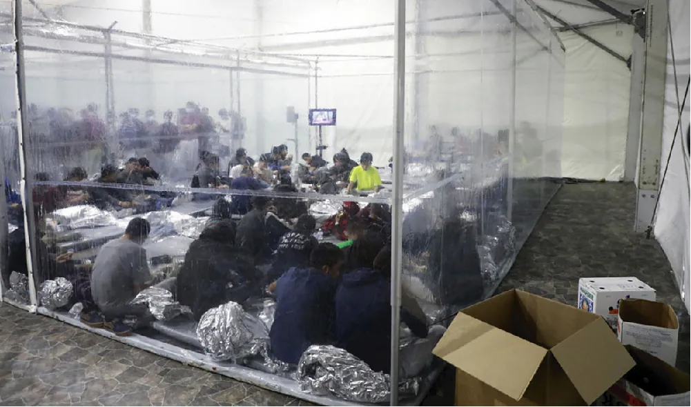 About twenty young people sit on the floor of a holding area with plastic sheeting as walls. Plastic blankets litter the floo