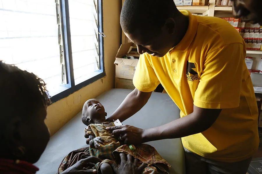 A person holds a baby who appears to be about two years old. The baby is on an examination table. In the background are boxes