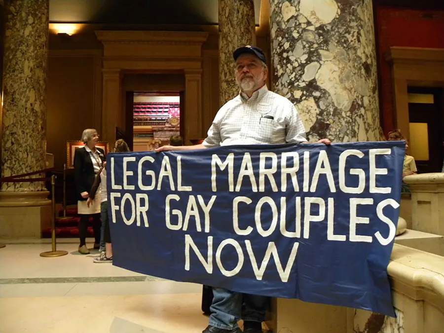 An older man with a gray beard, wearing a baseball cap, buttoned-up shirt, and jeans, is shown in a marbled lobby with column