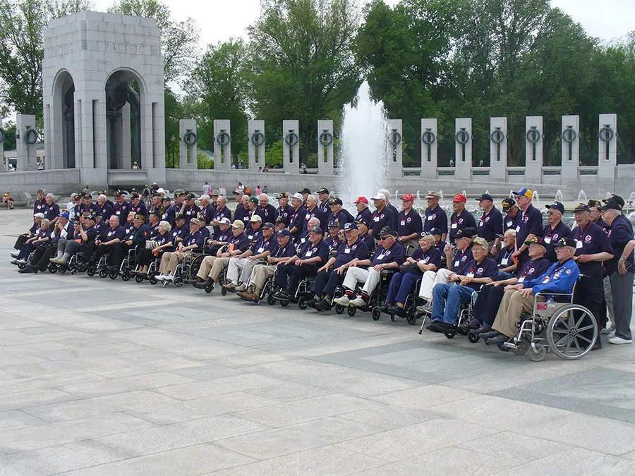 A group of elderly men, many in wheelchairs, all dressed in blue shirts and baseball caps, are shown standing and sitting in 