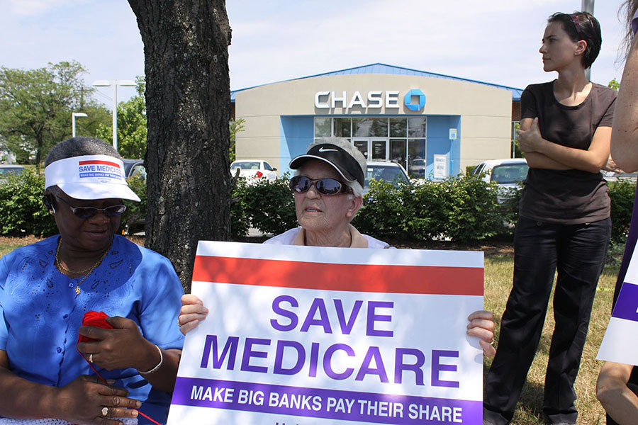 Two elderly women, one holding a red white and blue sign reading "Save Medicare: Make Big Banks Pay Their Share," are shown s