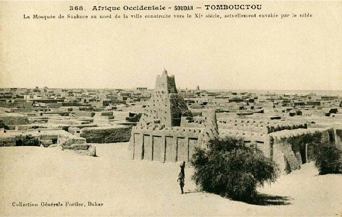 The postcard shows a brown, sandy mosque with tall walls and a seven-story building in the middle showing above the walls. No