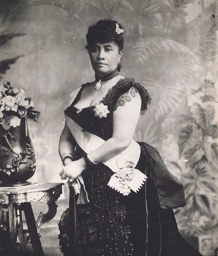 A woman is shown in a black and white photograph. She is wearing a low cut beaded dark dress with lacy cap sleeves. A brooch 