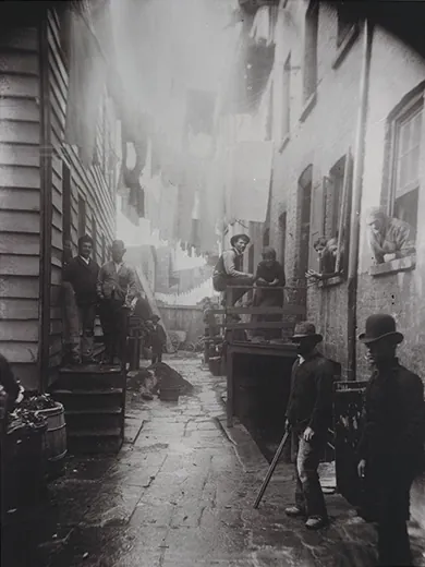 A photograph shows an alley between two tenements. Men, women, and children stand on either side of the alley, in the stoops,