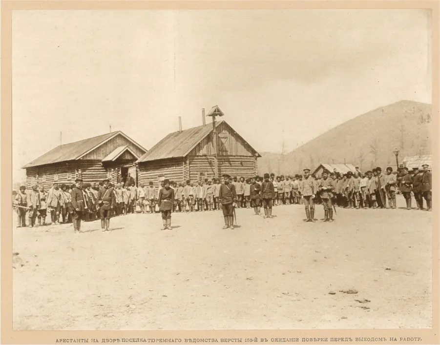 This photograph shows a group of workers standing in a line. Men in military style uniforms stand near the workers. Several l