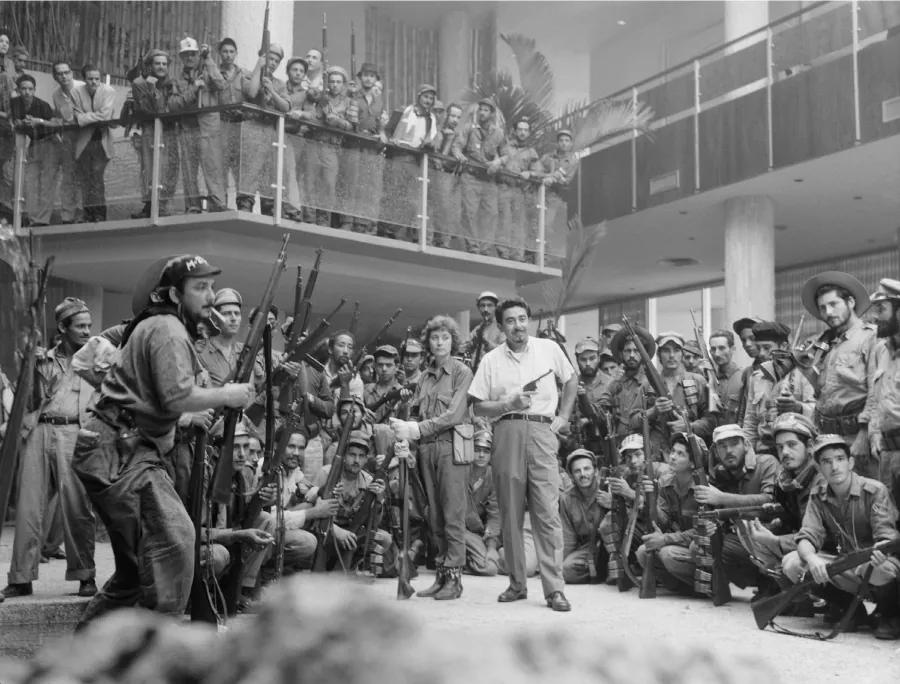 A photograph shows a man standing inside a building in a white short sleeved shirt and pants, holding a pistol in his right h