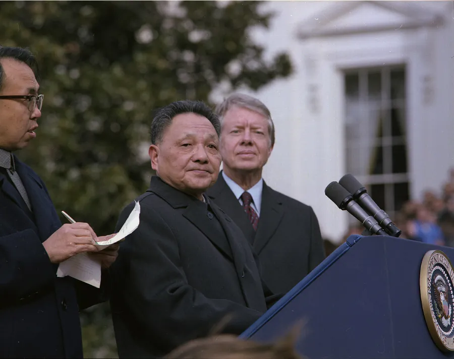 A photograph shows three men standing outside behind a podium with microphones. The man on the far left holds a paper and pen