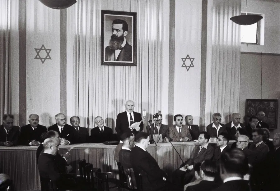 A photograph shows several men sitting behind a long table. A man in the middle stands, holds a piece of paper, and speaks in