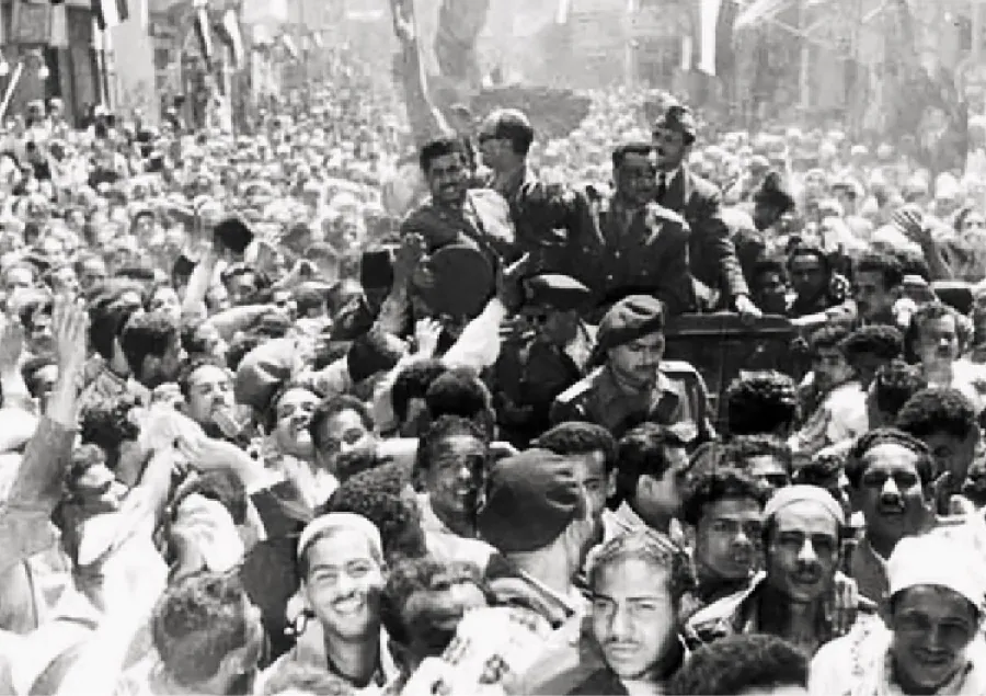 A photograph shows a crowd, mostly of men. In the middle of the crowd, seven men are riding in a car looking at the crowd. Th