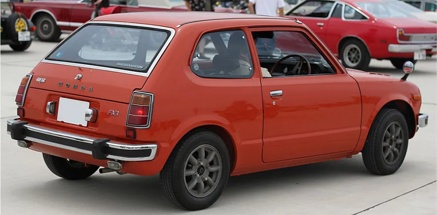 A picture of the side of a small hatchback, two-door, orange car is shown on white pavement. The car has black wheels with gr